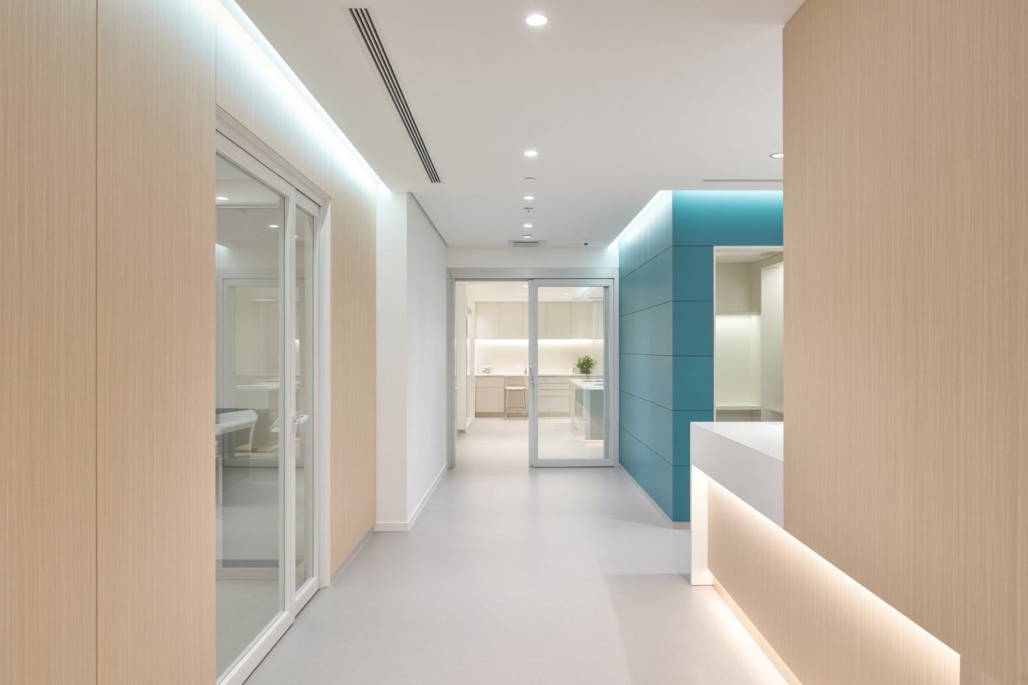 A bright, quiet surgery-center hallway with pale wood walls and a teal accent wall.
